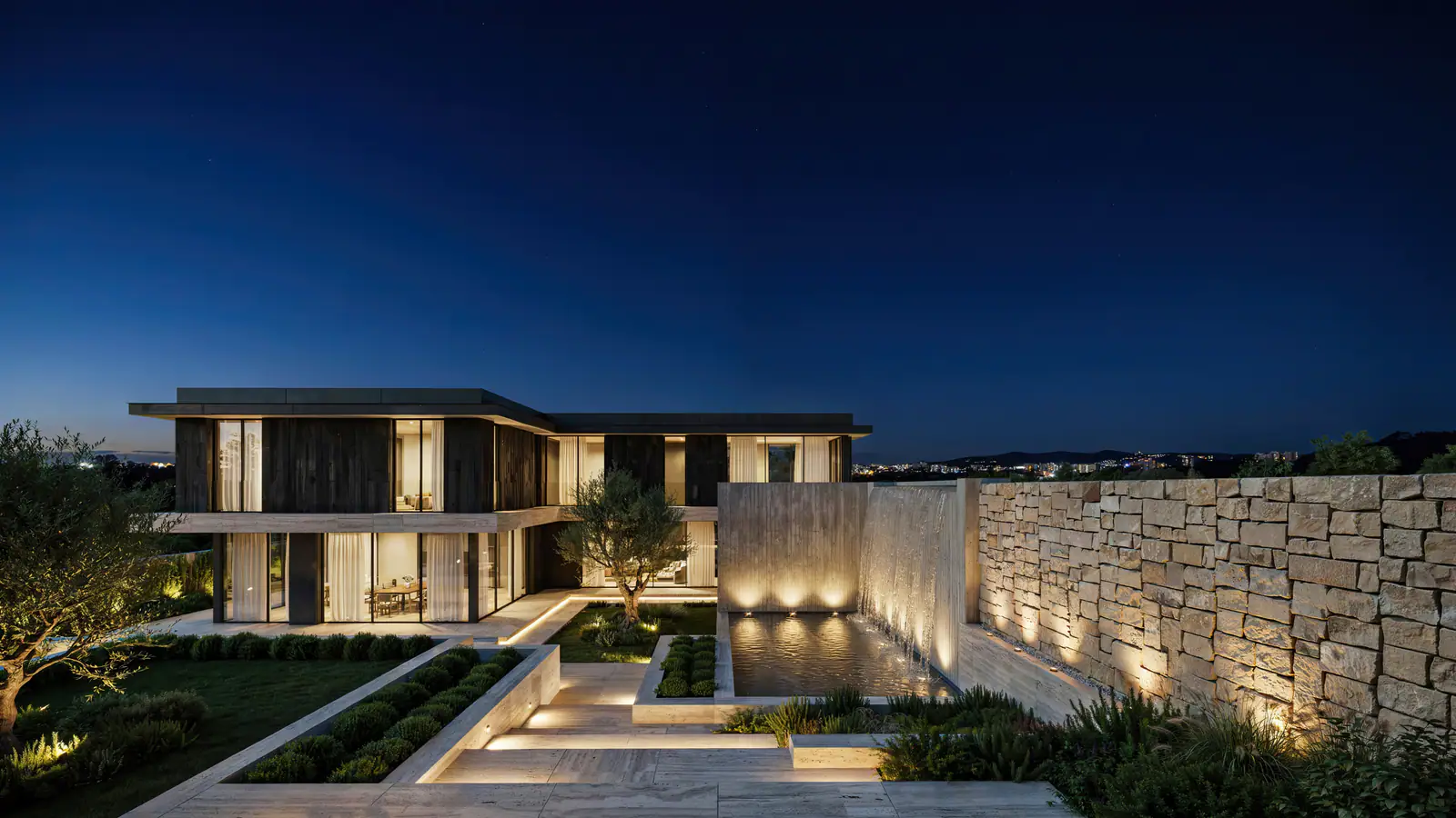 Modern glass residence beside a softly lit stone wall at blue hour
