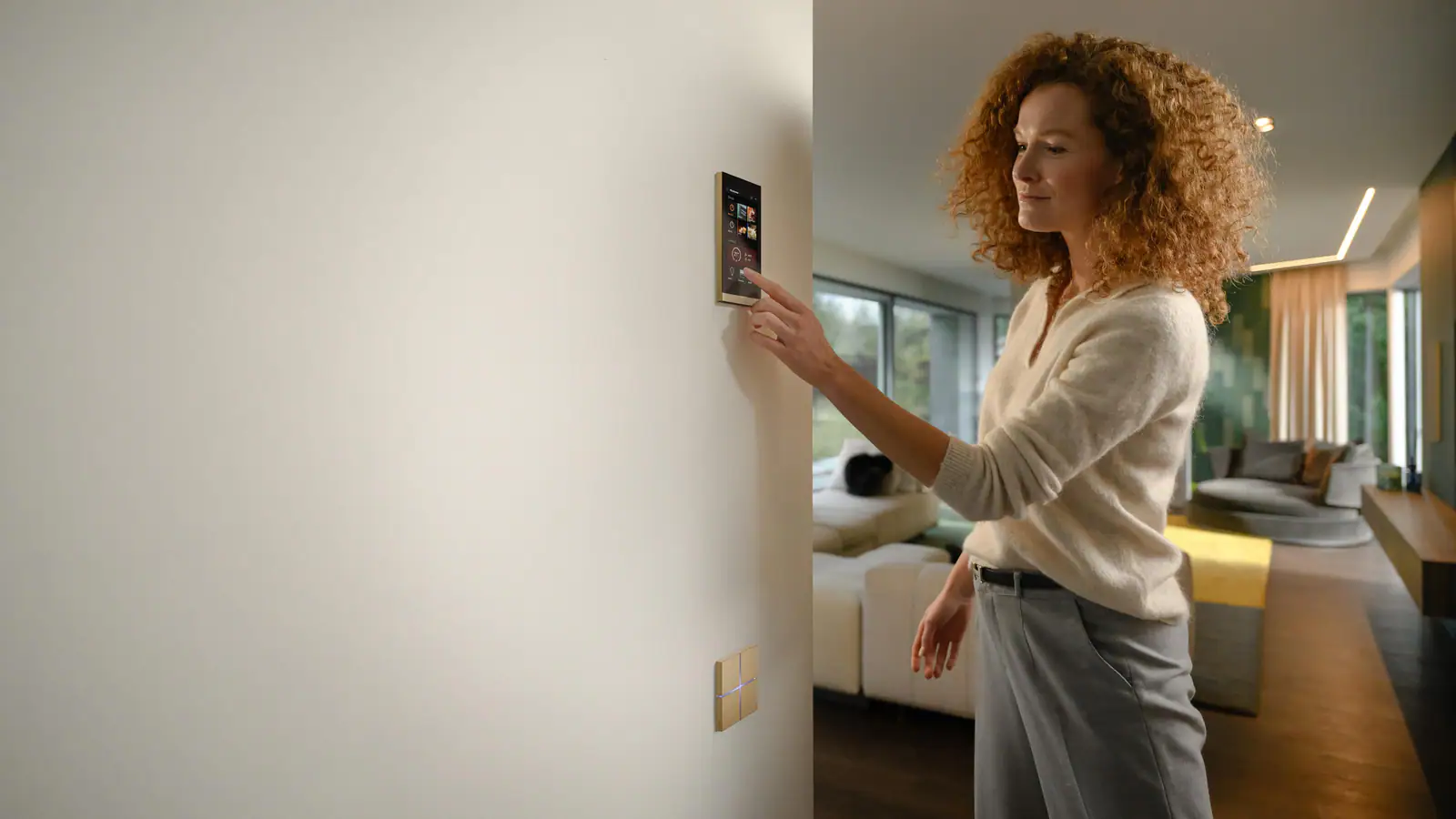 A woman touching a Basalte Ellie touch display, a brass Sentido keypad below it, with a living room and garden windows beyond