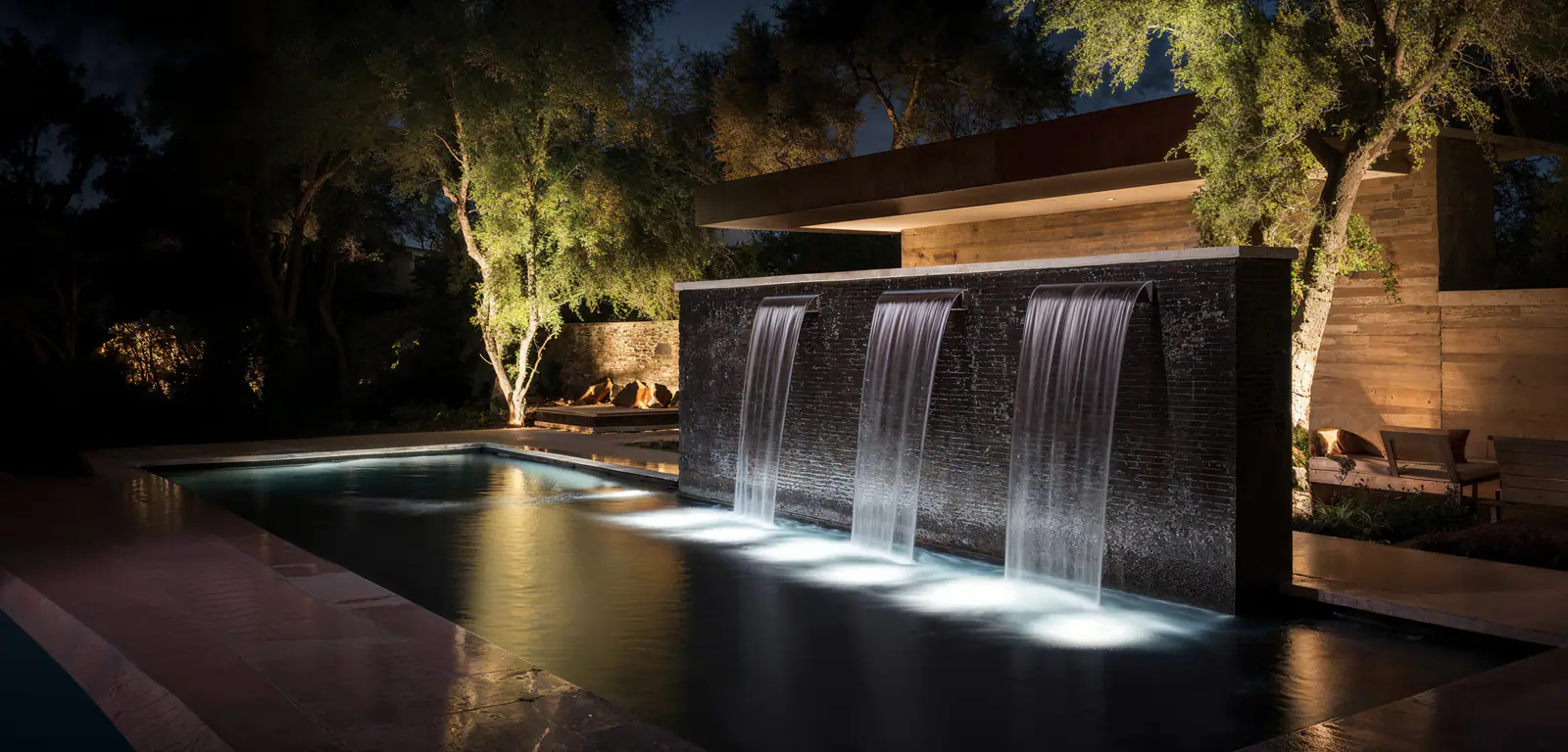 A lit water wall spilling into a pool at night, framed by softly uplit trees