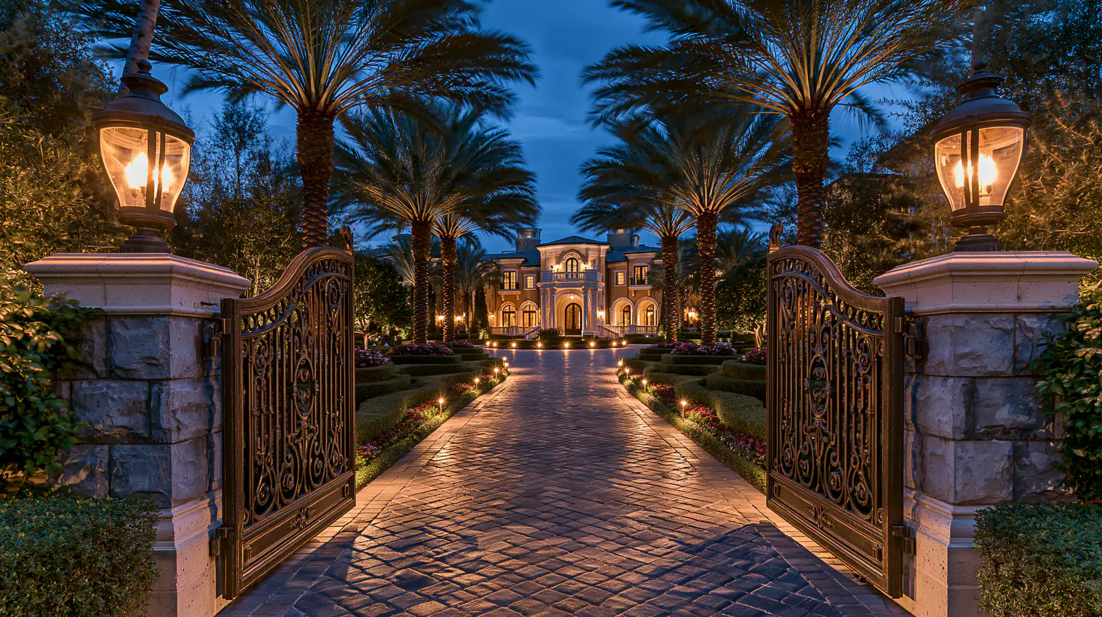 Estate gates opening onto a lantern-lit, palm-lined drive at night